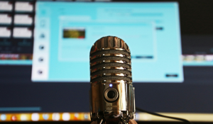 Close-up of a vintage-style podcast microphone with a glowing blue indicator light, set in front of a computer screen displaying a blurred user interface.